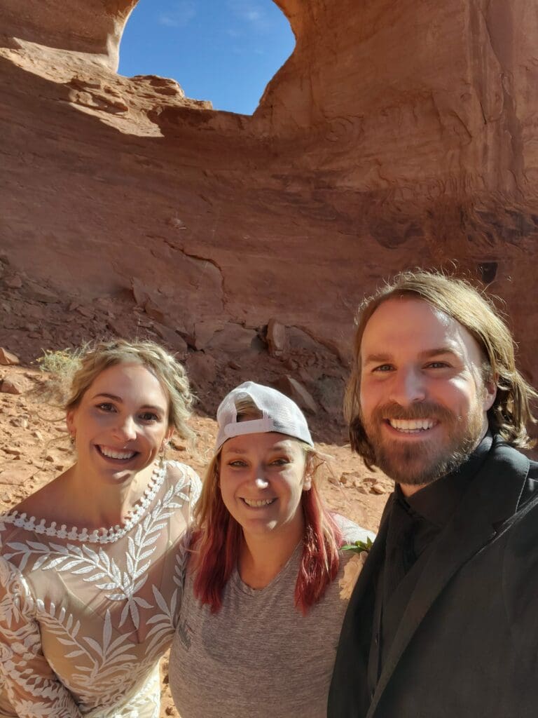 Elopement couple posing at Looking Glass Arch in Moab, Utah, accompanied by a photographer, with red rock desert scenery in the background.