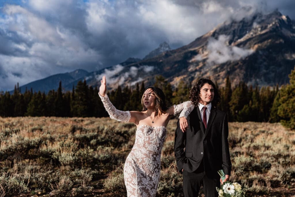 Elopement couple striking a romantic editorial pose at Mountain View Turnout with Grand Teton mountains and alpine meadow behind them.