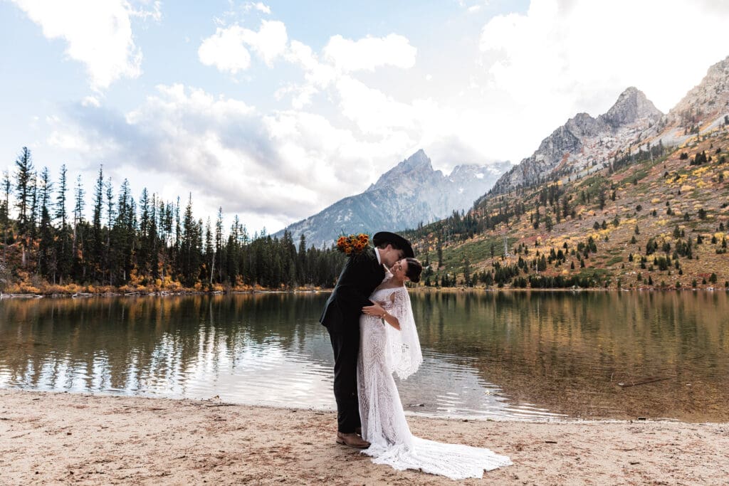 Elopement couple embracing by the calm waters of String Lake, with snow-capped Grand Teton peaks and evergreen trees reflected in the lake.