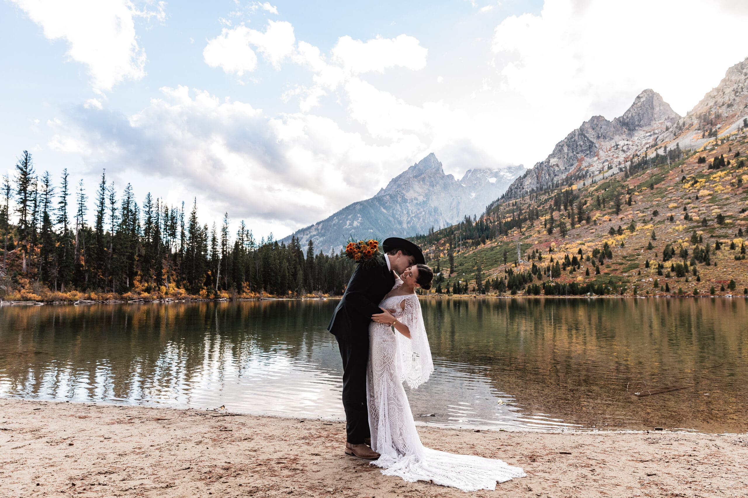 Elopement couple embracing by the calm waters of String Lake, with snow-capped Grand Teton peaks and evergreen trees reflected in the lake.
