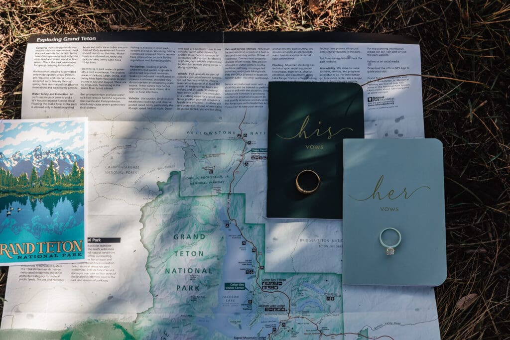 Flat lay of elopement details including wedding rings, vow books, and a map photographed at Schwabacher’s Landing in Grand Teton National Park.