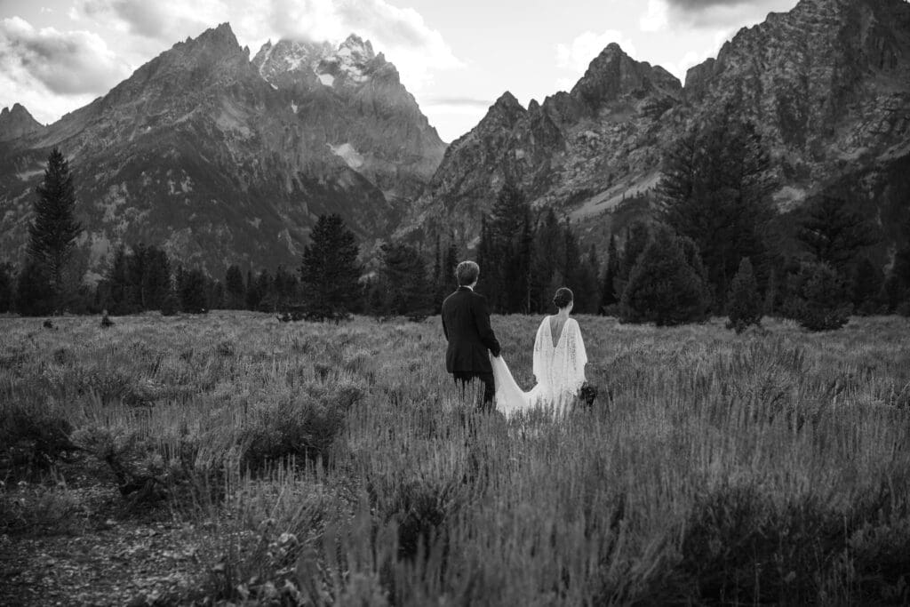 Groom holds the bride’s train as they walk hand in hand through vibrant alpine meadows with snow-capped peaks in Grand Teton National Park.
