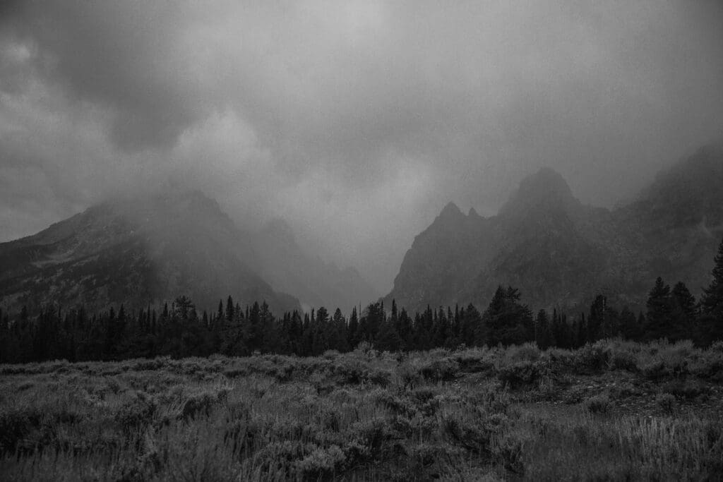 Black and white landscape of misty mountain peaks rising above forest and meadow during moody weather in Grand Teton National Park.