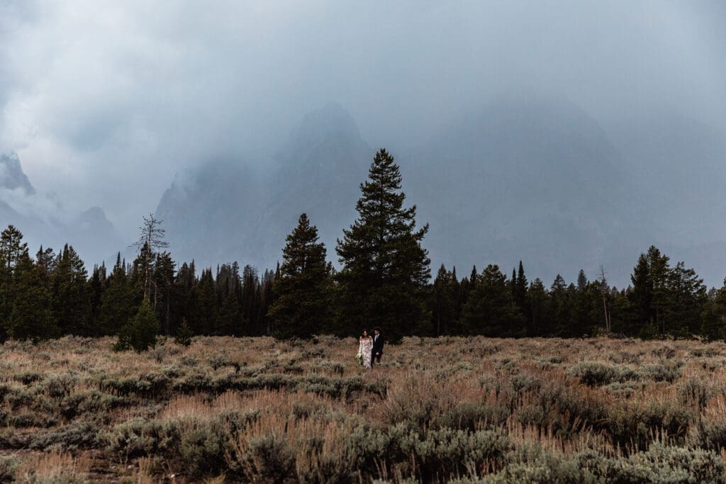 Elopement couple walking hand-in-hand through Mountain View Turnout, surrounded by vibrant fall foliage and dramatic Grand Teton peaks at sunrise.