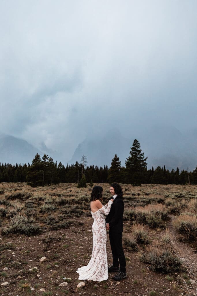 Elopement couple in wedding attire standing in a scenic Grand Teton National Park landscape with moody clouds and evergreen trees.
