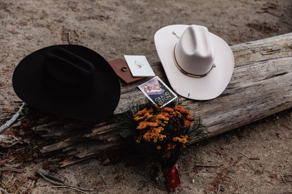 Two cowboy hats placed beside orange flowers on a log at String Lake during a Grand Teton National Park elopement.