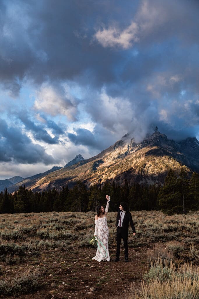 Elopement couple celebrating their marriage at Cascade Canyon Turnout with dramatic storm clouds and golden evening light in Grand Teton National Park.