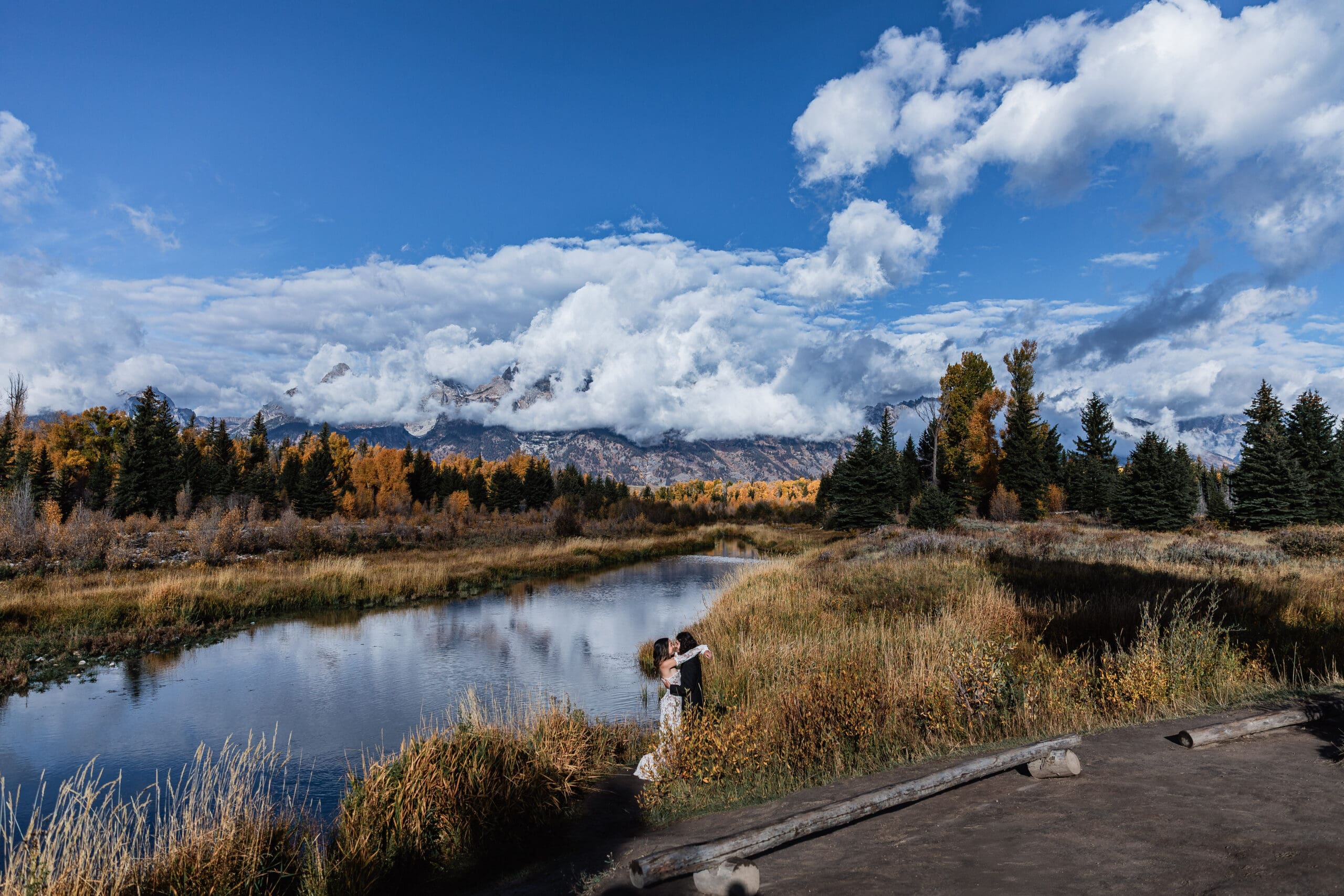 Elopement couple hugging and taking in the Grand Teton mountains reflected in the calm lake at Schwabacher Landing, surrounded by autumn trees and golden reeds.