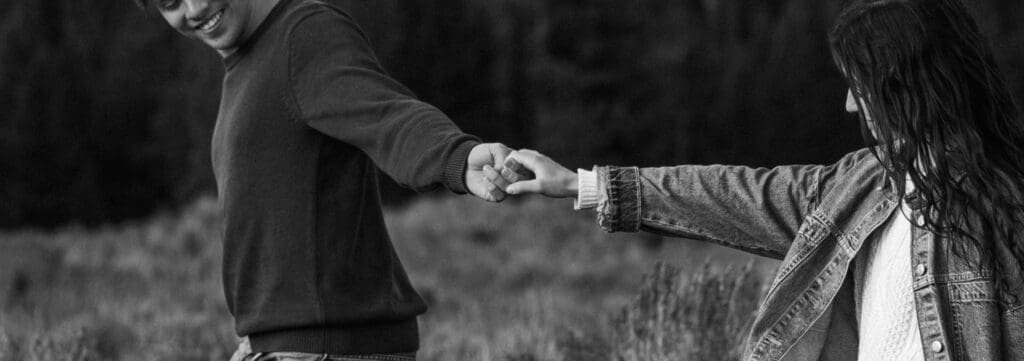 Black and white engagement photo of a couple holding hands and leaning away from each other in an open field at Grand Teton National Park.