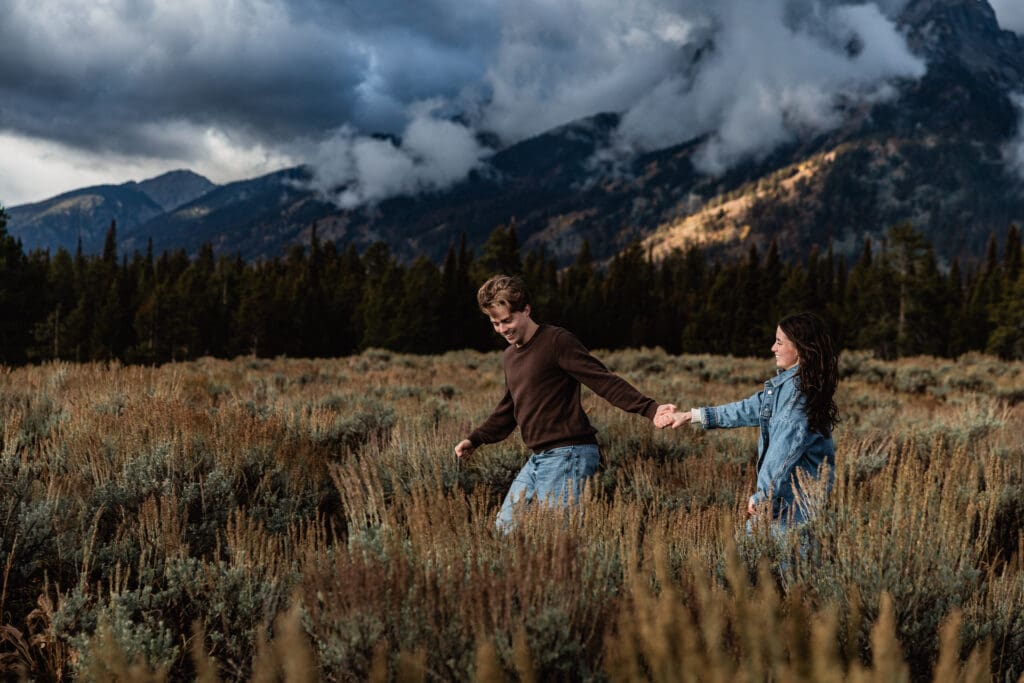 Dramatic mountain landscape with storm clouds as people walk through golden grass fields in Grand Teton National Park.