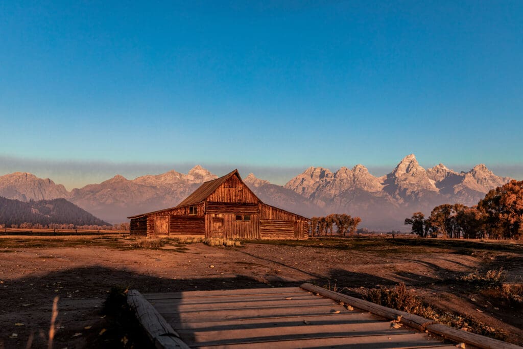 Rustic wooden barn at Mormon Row with snow-capped Grand Teton peaks glowing in warm sunset light in Grand Teton National Park.