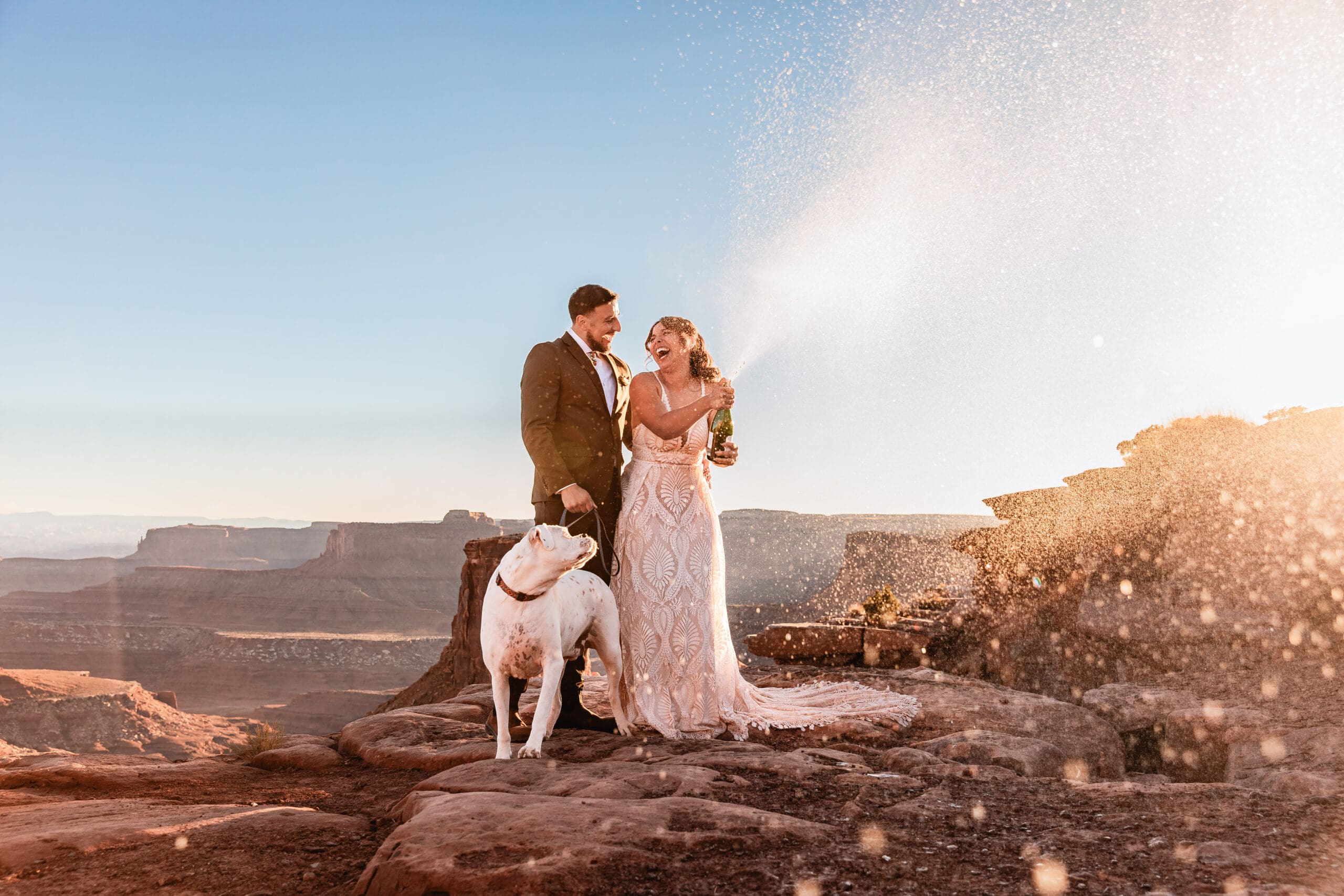 Elopement couple celebrating with a champagne pop at Marlboro Point in Moab, while their dog steals the moment with the best reaction during their desert wedding adventure.