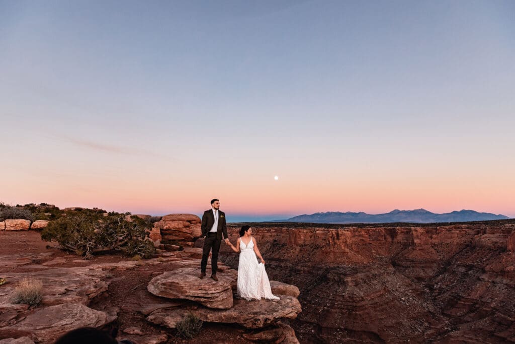 Groom standing on a rock above the bride, holding hands under the rising moon at Malboro Point, Moab.
