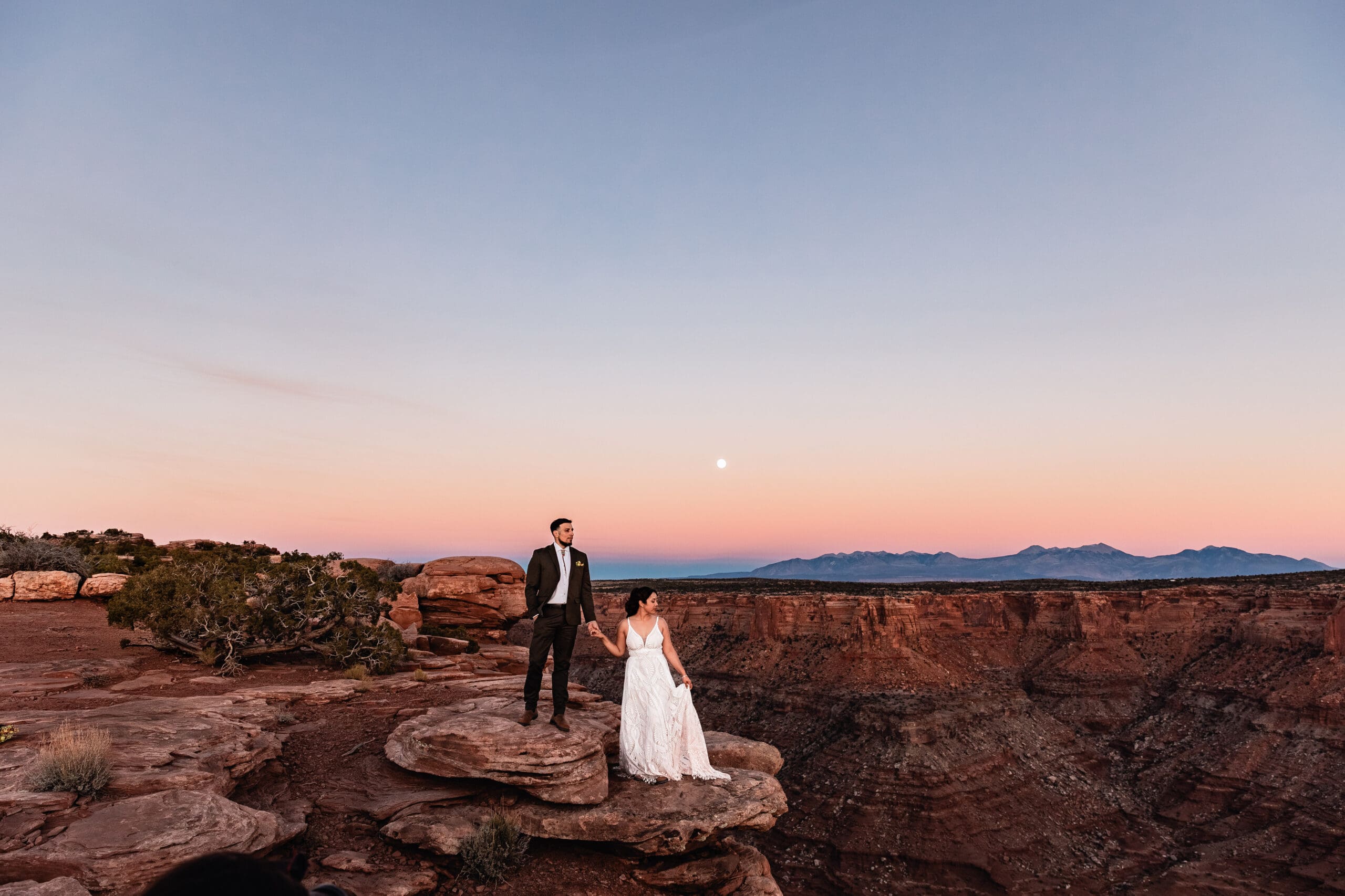 Groom standing on a rock above the bride, holding hands under the rising moon at Malboro Point, Moab.