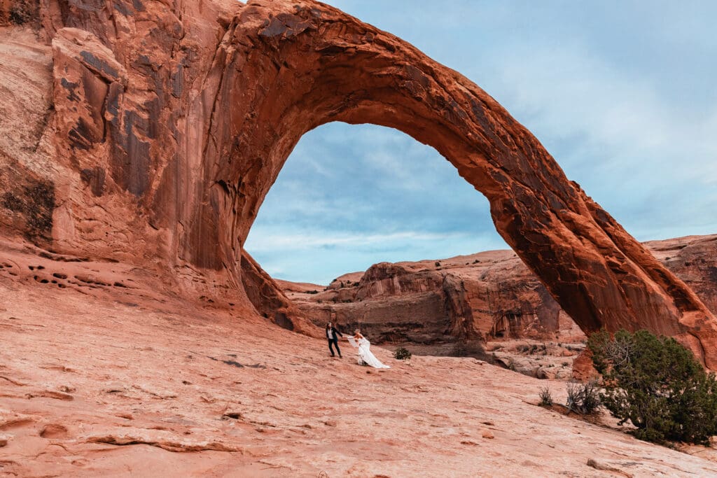 Elopement couple run under Corona Arch in Moab, Utah, set against bold red sandstone formations and a vivid blue desert sky.