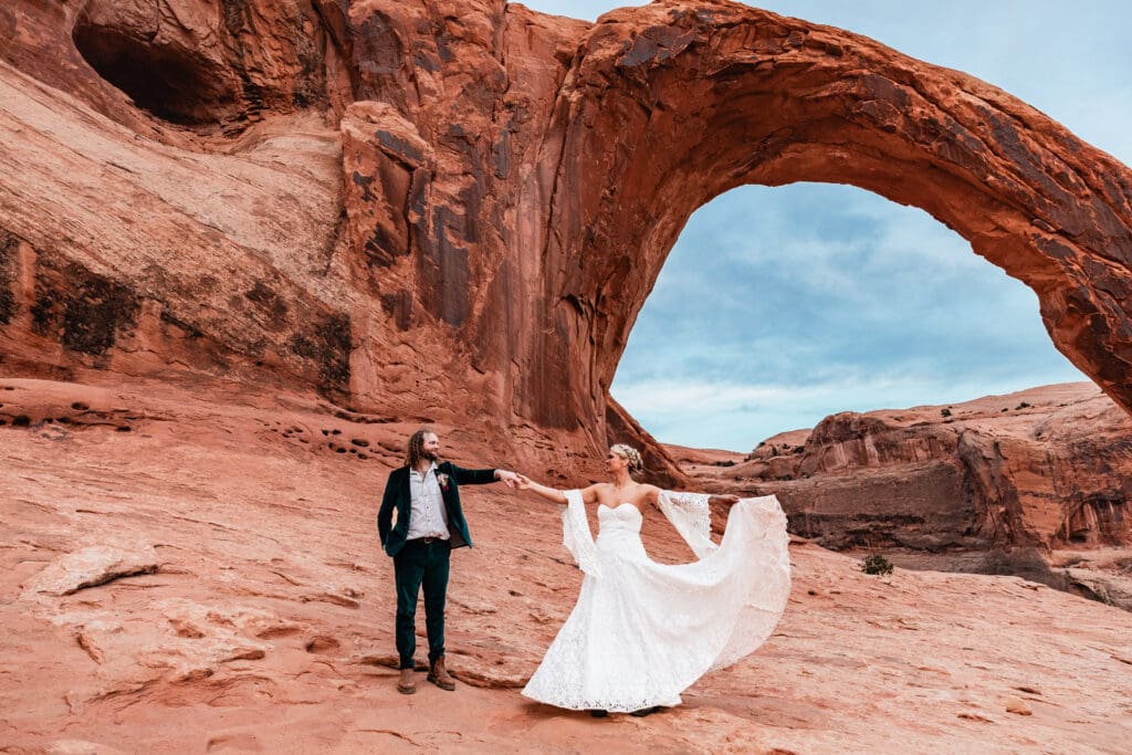 Bride twirling in a dramatic spin while groom watches, capturing joyful movement during their elopement at Corona Arch, Moab.