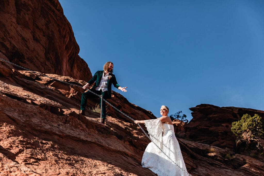 Bride and groom in wedding attire climbing the chain section along Corona Arch trail, capturing adventure and romance in Moab.