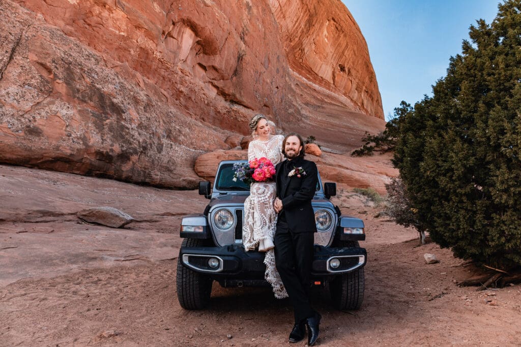 Bride and groom sitting on the Jeep hood, looking in different directions while enjoying their Moab elopement adventure near Looking Glass Arch.