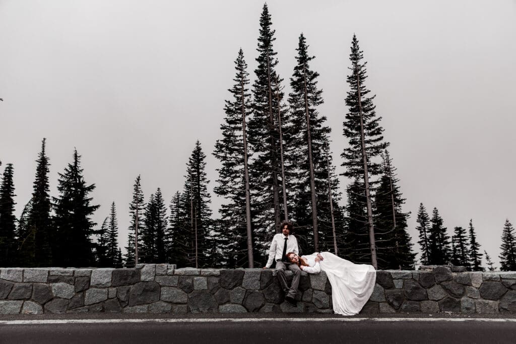 Elopement couple sitting on a stone wall with tall evergreen trees silhouetted in the background in Mt. Rainier National Park.