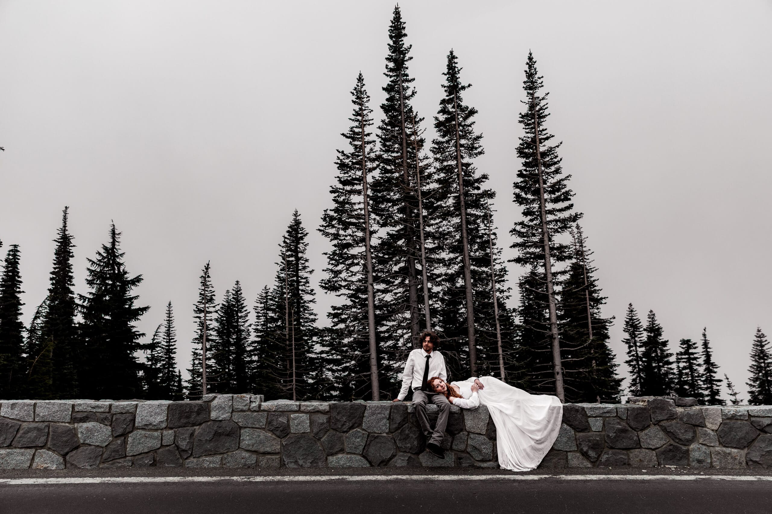Elopement couple sitting on a stone wall with tall evergreen trees silhouetted in the background in Mt. Rainier National Park.