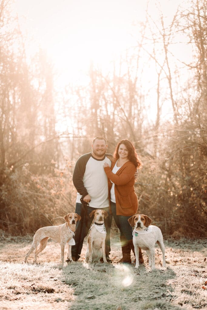 Couple stands outdoors in warm sunlight with their three dogs on a frosty morning in the woods during an engagement session