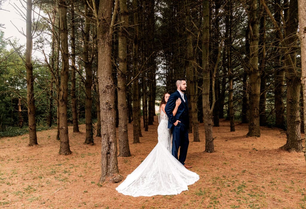 Bride wrapping her arms around her groom from behind while standing in the pine trees at Sanaview Farm wedding in Pennsylvania