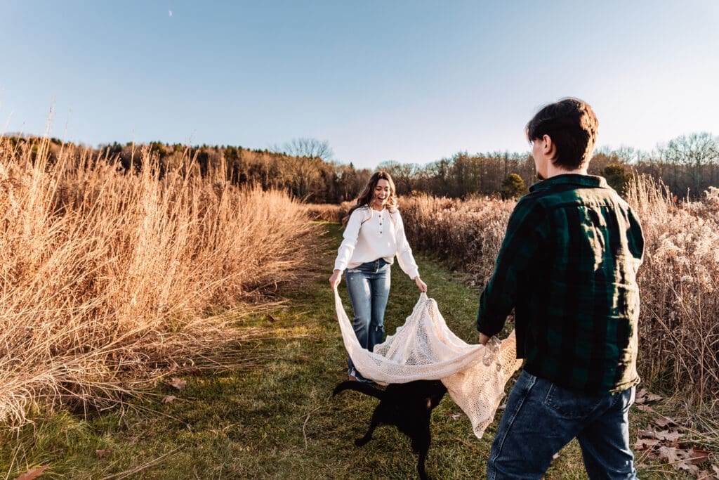 Couple plays with their dog on a grassy path surrounded by tall dried cattails and brush during a sunny autumn engagement session