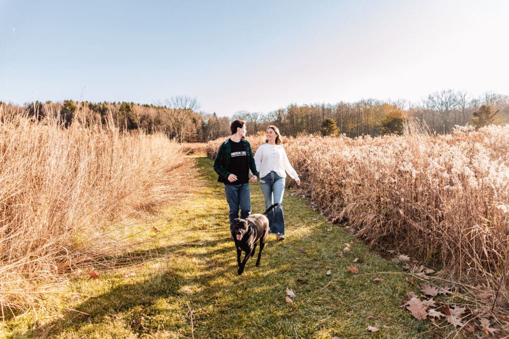 Couple runs together with their dog on a grassy path surrounded by tall autumn grasses during a sunny engagement session