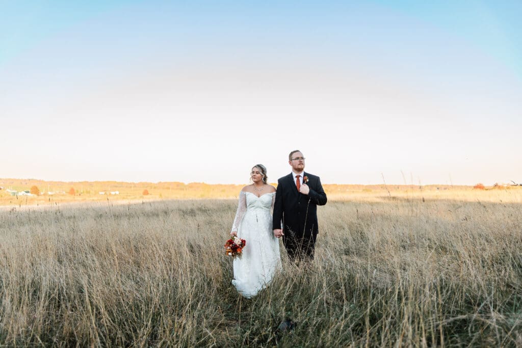 Newlywed couple stand together through golden prairie grass field at sunset after their Willowbrook ceremony