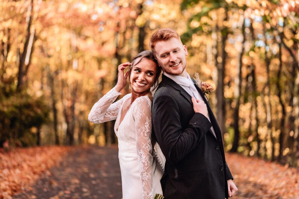 Wedding couple poses together on a driveway lined with vibrant fall foliage at The Barn At Ever Thine