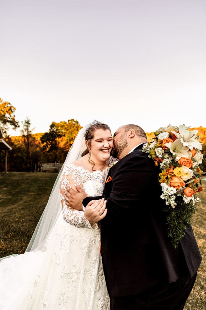 Groom kisses his bride’s face as she laughs during portraits at fall Ridgemont Farm wedding in Beaver County