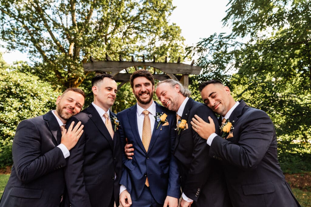 Groomsmen in navy suits sharing candid laughing moments outdoors at Succop Nature Park wedding