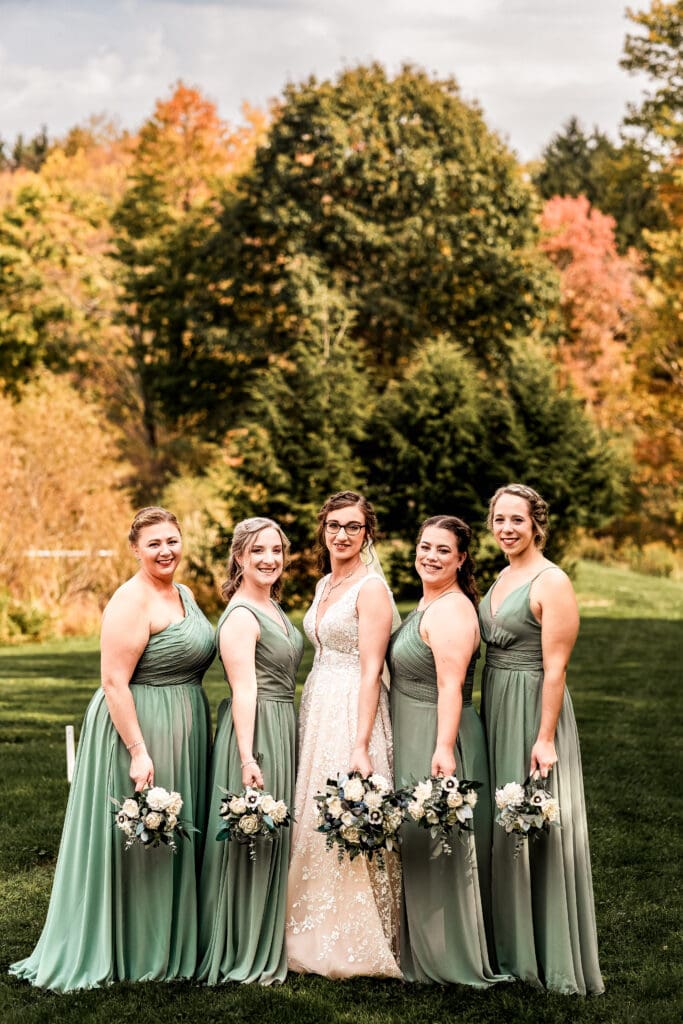 Bride with bridesmaids in sage green dresses with fall foliage and pond in background at Pinehall at Eisler Farm