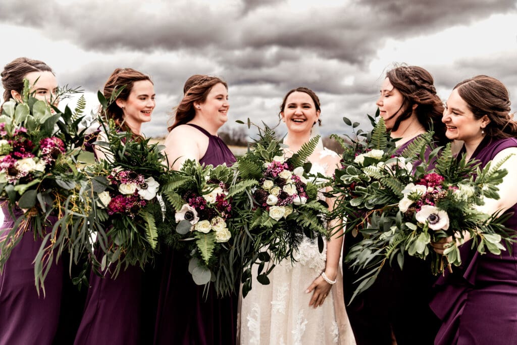 Bride and bridesmaids laughing together while holding lush bouquets during an outdoor Lingrow Farm wedding