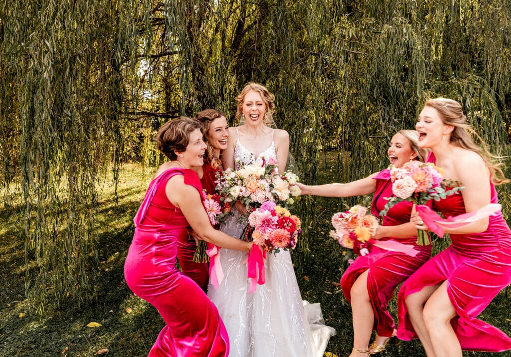 Bridesmaids running to hug the bride under a willow tree at The Gathering Place at Darlington Lake