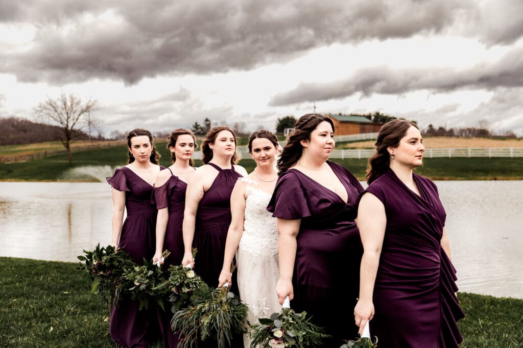 Bride and bridesmaids in deep purple dresses posing by a pond under moody skies at a Lingrow Farm wedding