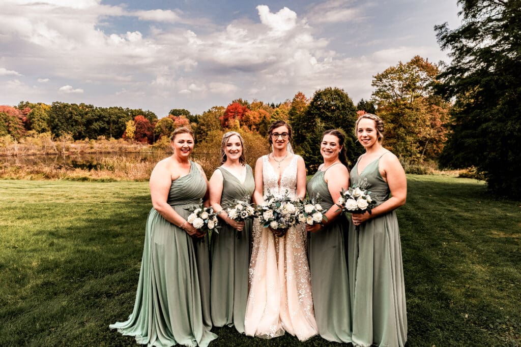 Bride and bridesmaids in sage green dresses posing in front of fall foliage and pond at Pinehall at Eisler Farm