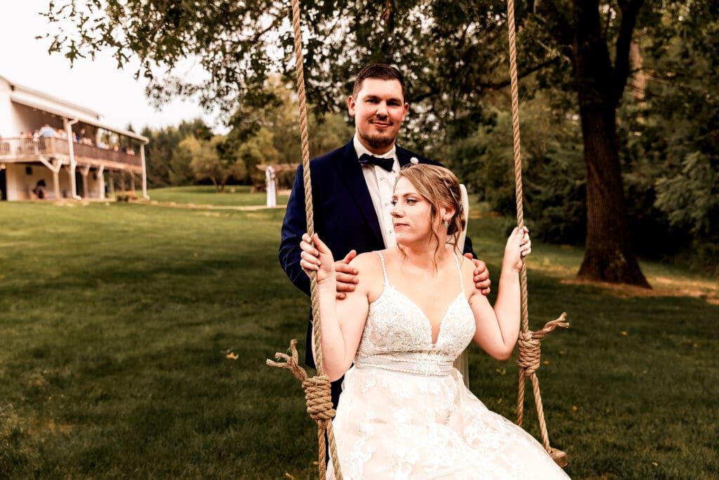 Bride sitting on a swing and looking into the distance while groom smiles during portraits at The Barn at Ever Thine in Fenelton, PA