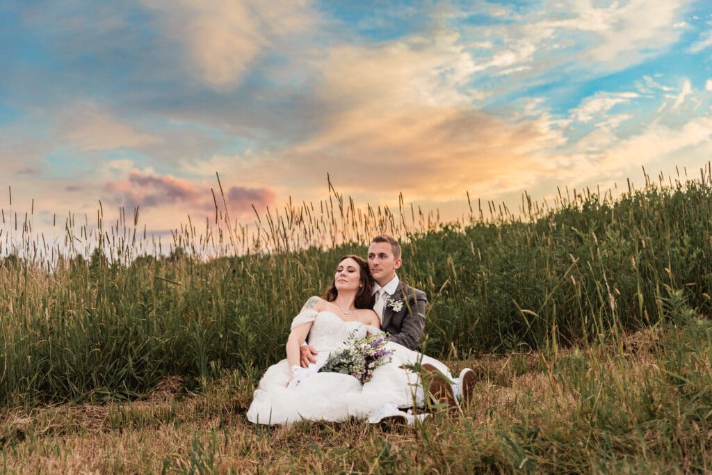 Wedding couple sits together in a grassy field at sunset with dramatic sky at Lingrow Farm