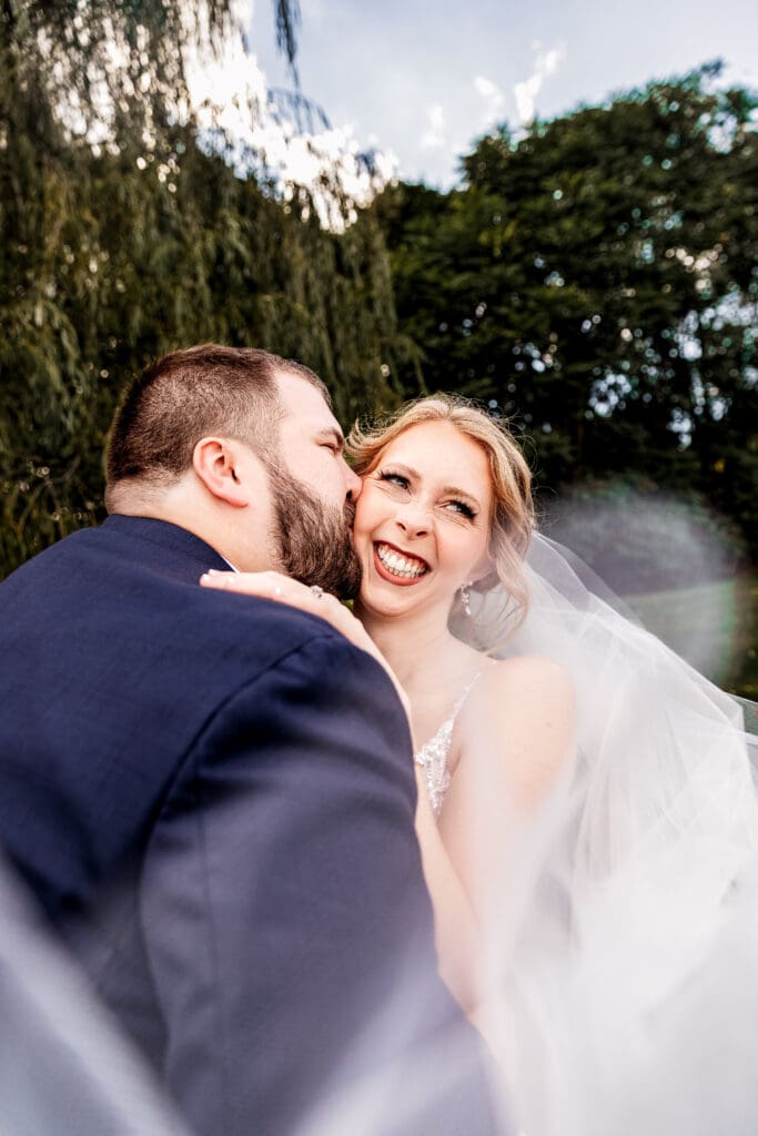 Groom kissing bride on the cheek under willow tree during portraits