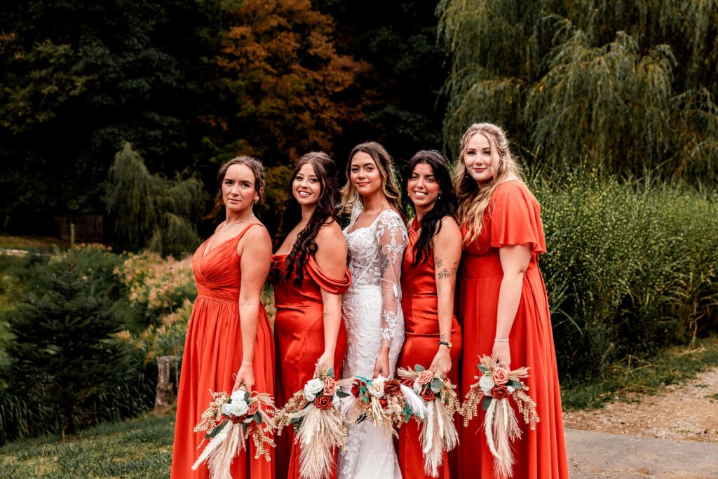 Bride and bridesmaids in terracotta and white dresses posing with willow trees and fall foliage at Hinckston Run Farm