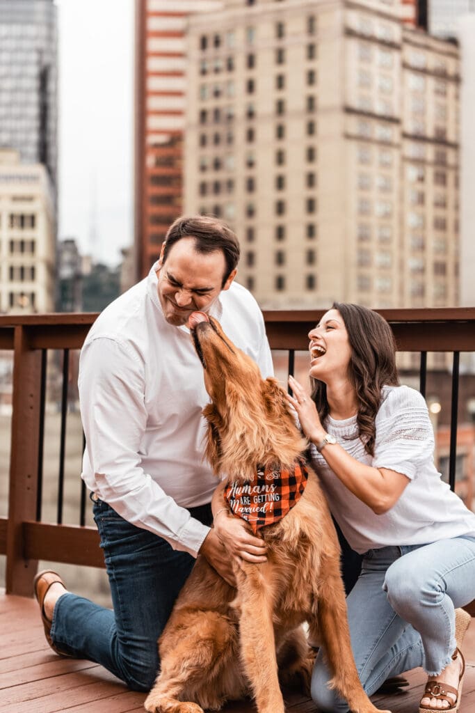 Couple plays with their golden retriever on an urban rooftop deck with city buildings in the background during an engagement session