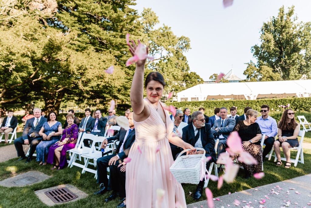 Adult flower girl playfully tossing flower petals toward the camera at a Pittsburgh Phipps Botanical Gardens wedding