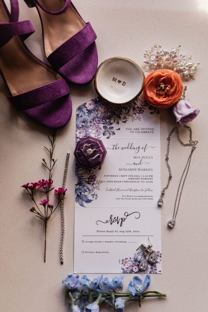 Wedding detail flat lay featuring purple heels, personalized ring dish, and orange florals at Pittsburgh Airport Marriott