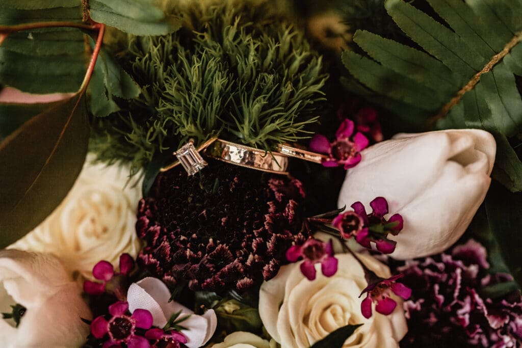 Close-up flat lay of gold wedding bands and emerald-cut diamond ring nestled in a moody floral arrangement with roses, greenery, and tulips at Lingrow Farm
