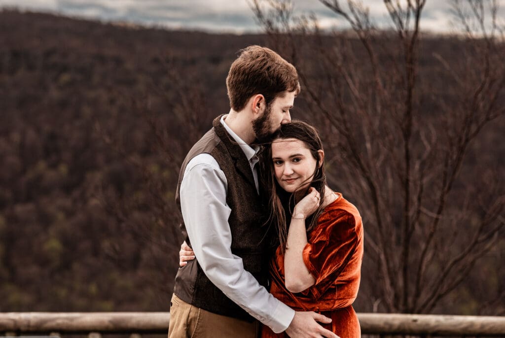 Bride-to-be holding her hair back as her fiancé kisses her forehead during a Coopers Rock engagement session