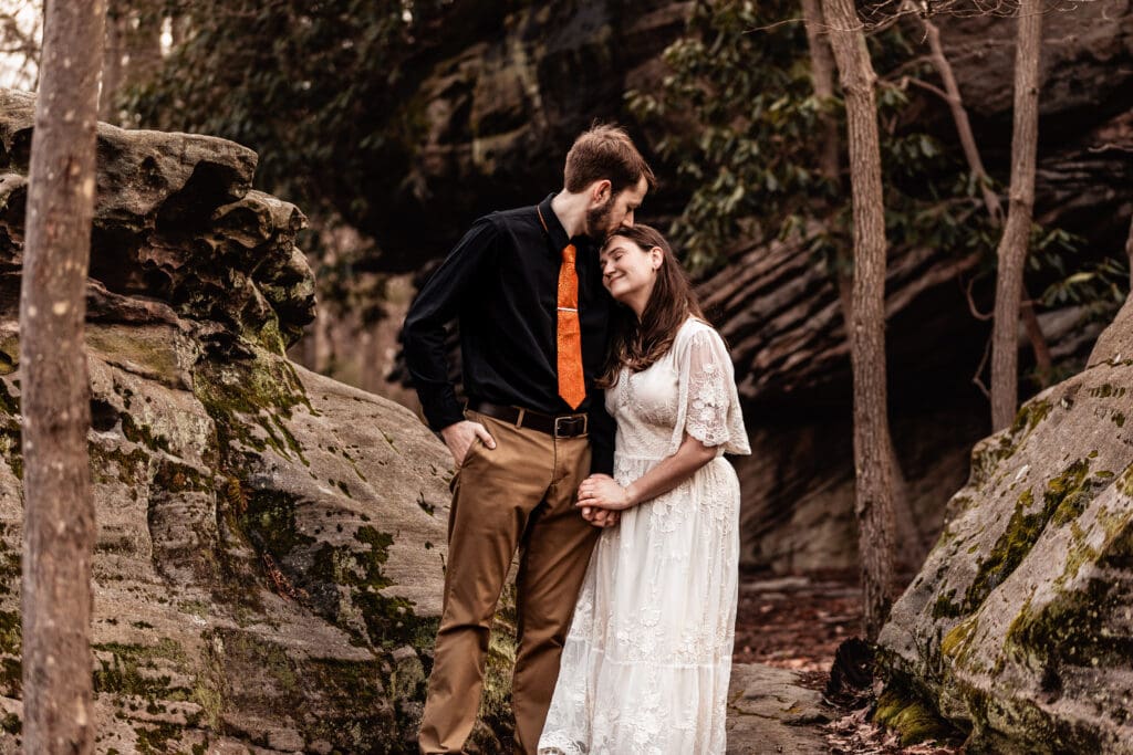 Bride-to-be resting her head on her fiancé’s shoulder as he kisses her forehead in the woods at Coopers Rock