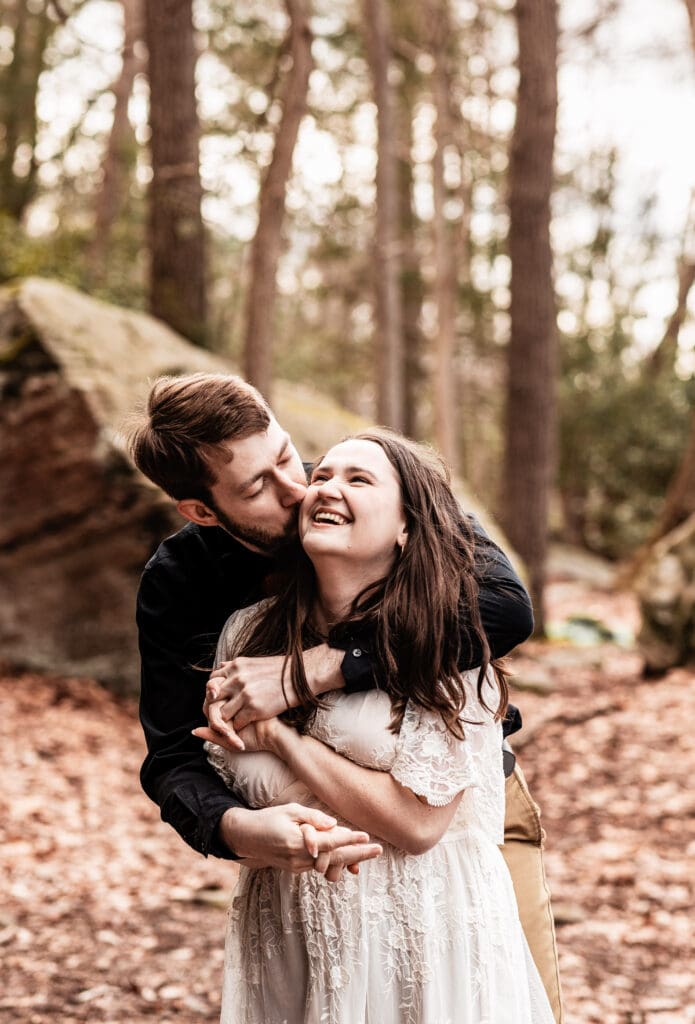 Groom hugging his fiancée from behind and kissing her cheek during a Coopers Rock engagement session