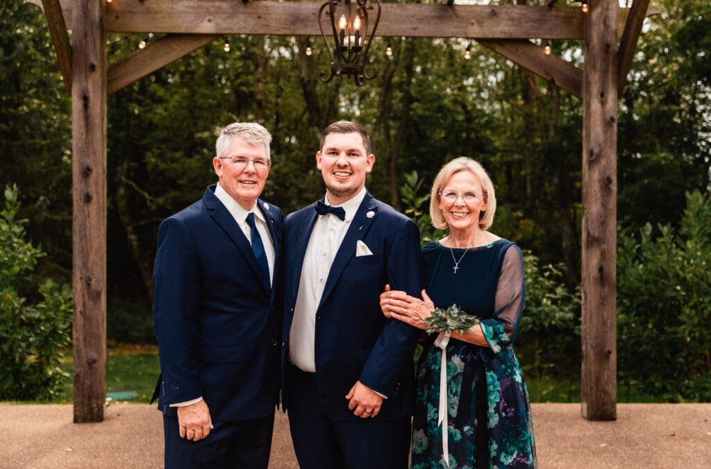 Groom poses with his parents under a wooden pergola at The Barn at Ever Thine in Butler County, Pennsylvania