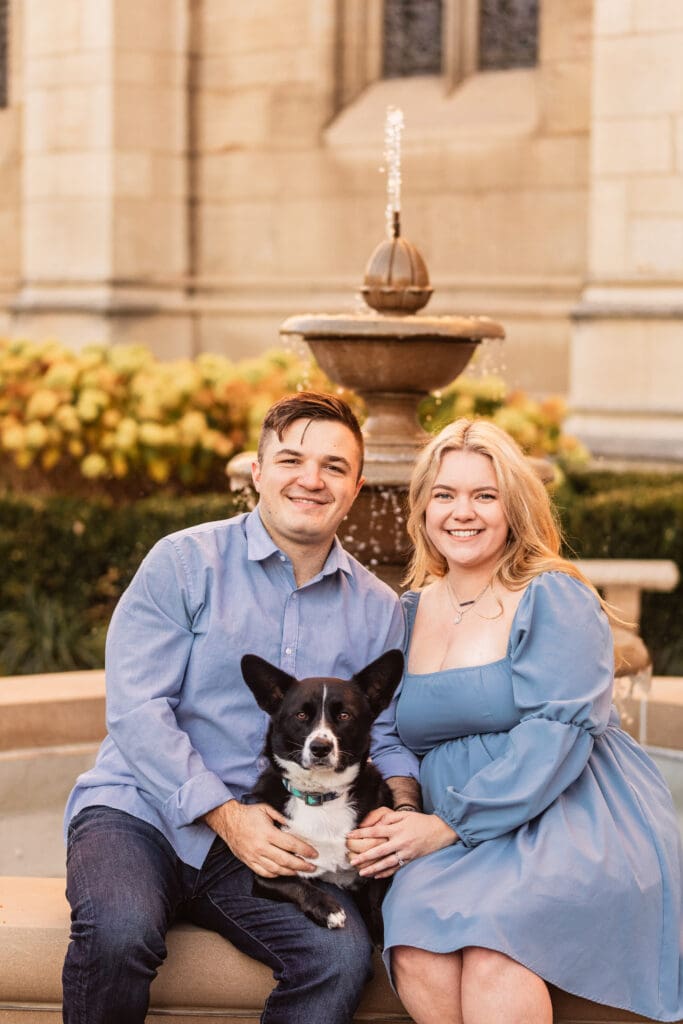 Couple wearing coordinating blue outfits sits outdoors on stone steps with their black and white corgi during an engagement session